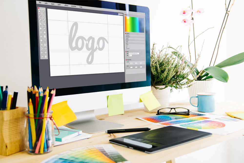 Schreibtisch mit Grafiktablet, Stiften und Tasse. Bildschirm mit geöffnetem Grafikprogramm und dem Wort "Logo" in der Mitte. Erarbeitung eines Styleguide.