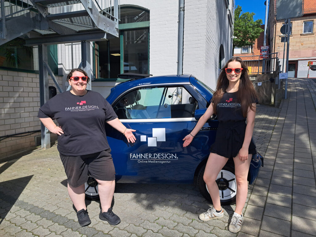 Zwei Frauen stehen neben einem blauen Auto mit dem Logo von Fahner.Design. Beide tragen schwarze T-Shirts mit dem Firmenlogo und rote Sonnenbrillen. Sie lächeln und zeigen auf das Auto.