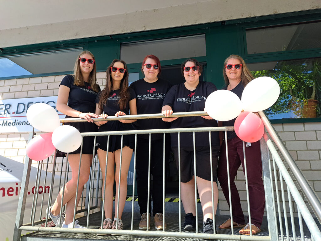 Fünf Frauen stehen auf einer Treppe vor einem Gebäude, alle tragen schwarze T-Shirts mit dem Logo von Fahner.Design und rote Sonnenbrillen. Zwei Luftballons sind an das Geländer gebunden. Sie lächeln und schauen in die Kamera.