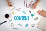 Weißer Tisch mit ‚Content‘, Lupe, Kopfhörer, Glühbirne und Handy, zwei Personen schreiben in Block.