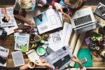 Vier Frauen sitzen an überfülltem Tisch mit Magazinen und Computer, Blick von oben.
