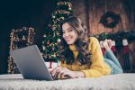 Frau liegt lachend auf einem Teppich vor dem Computer, im Hintergrund Weihnachtsbaum und Dekoration