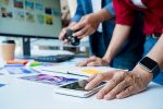 Tisch mit Handy und Bildern, im Hintergrund Computer und eine Fotokamera.