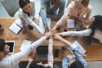 Ein Team aus sieben Menschen die an einem Tisch zusammensetzen und ihren rechten Arme in die Mitte des Tisches halten